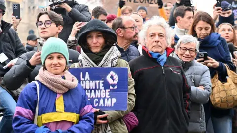 Reuters A group of people stand facing the camera. One at the front is colourfully dressed, and next to her a woman holds a sign in French that reads: Shame changes sides