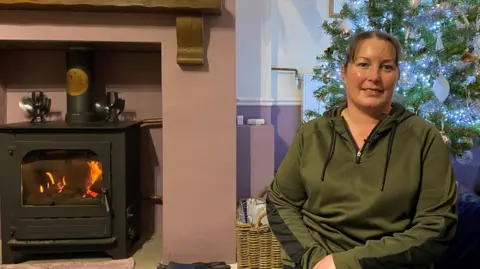 BBC Sam Wood is sitting in front of a Christmas tree with a fire to her left. She's wearing a green top and has her hair tied back 