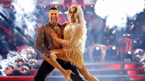 BBC Studios/Guy Levy Tayce dancing with Kai Widdrington on Strictly. Tayce is wearing a short gold fitted dress and has long blonde hair. Kai is wearing a sheer black and silver shirt and black trousers and there is a Christmas tree in the background