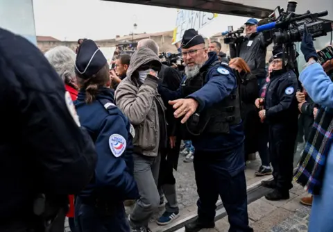 Getty Images One of the Pelicot case defendants wearing a face mask, hooded jacket and sunglasses arrives at the court house surrounded by cameras and police.