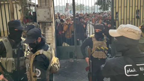 Aamir Peerzada/BBC Armed and masked men stand inside a gate, while a crowd of other men press against the gate.