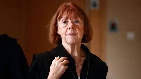 EPA Gisèle Pelicot, a woman with red hair in a black top, looks at the camera. She is not smiling