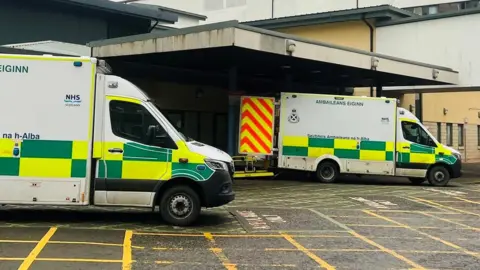 BBC Ambulances with green and yellow checked markings, parked outside a hospital building.