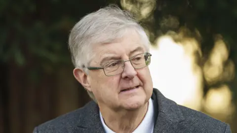 PA Media Mark Drakeford is wearing a grey suit and glasses in front of a blurred background