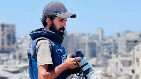 BBC A man with a beard, wearing a baseball cap, holds a professional-standard camera. He is in profile and wearing a flak jacket with the word "Press" on it, and he appears to be standing on the upper level of a building on a sunny day. In the background is the blurred image of what appears to be a heavily bomb-hit city.