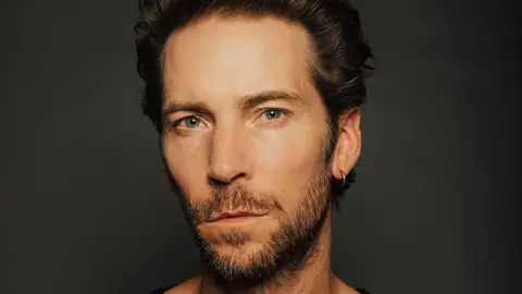 Heirlume An artistically lit headshot of a man in front of a dark background. He's got a neutral expression, a silver hoop earring in his left ear and his angular chin is framed by a short beard.