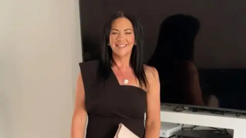 PSNI Karen Cummings, who has long dark hair parted in the middle, smiles while wearing a black dress and silver necklace and standing in front of a large flat screen TV 