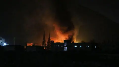 Getty Images At night, buildings are back-lit from a bright red light, which billows smoke into the air