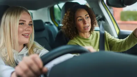 Getty Images A young woman with blonde hair is behind the wheel of a car. Beside her, a brown-haired woman indicates with her hand as she speaks