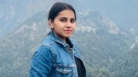 Northamptonshire Police Harshita Brella, who has tied-back black hair and is wearing a denim jacket and a black top. She is posing for a photo in a high spot with tree-covered mountains behind her.