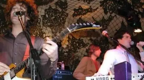 Chris Steele The Killers performing in a dark club in 2003. A man wearing a white shirt  sings into a microphone, while a guitarist with long curly hair sings along and a keyboardist plays in front of a wall decorated with a psychedelic design.