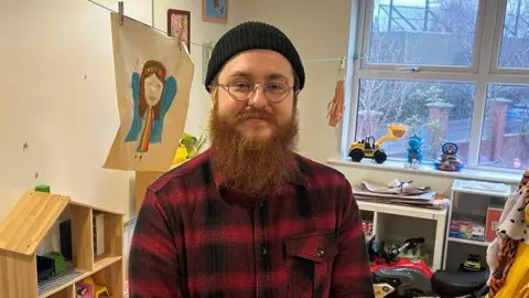 BBC Aaron pictured with a black beanie hat on and has a long ginger beard. He is wearing a black and red plaid shirt and thin gold rimmed circular framed glasses. 