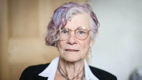 BBC Head and shoulders image of Nikki MacLeod, who was a victim of an online romance scam. She is 77, has thin-rimmed glasses, short grey hair with purple streaks and is looking straight at the camera. She has a dangly cross earring in her left ear and is wearing a beaded necklace.