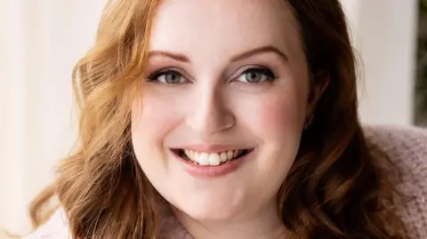 Elodie Guige Jen has long, wavy reddish-brown hair. She is wearing a pale pink knitted cardigan and is smiling at the camera.