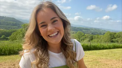 Heledd Roberts Heledd Roberts smiling as she looks at the camera. Behind her, you see rolling hills and a blue sky in what is a picturesque location. There are tree and green areas scattered around the background suggesting Heledd is in a rural location.