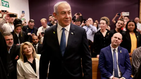 Reuters Netanyahu stands in a courtroom wearing a black suit jacket, a white shirt and a blue tie. Behind him the room is crowded, with several people holding their phones aloft to take photos of him. 