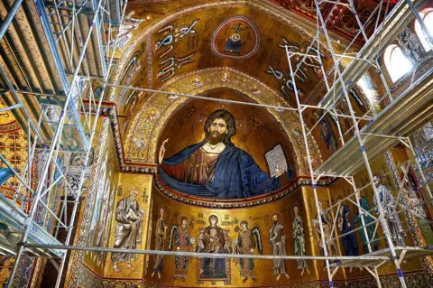 Reuters The apse of Monreale's cathedral in Sicily, showing a detailed mosaic of Christ and other religious symbology in mostly gold, with scaffolding in the foreground. 