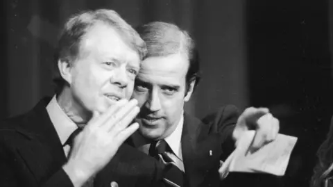 Getty Images Jimmy Carter, with a hand over his mouth as if to speak in a noisy room, leans towards Joe Biden who is pointing into the distance. Both men are wearing suits and appear to be on stage. The photo was taken at a fundraiser in Wilmington in 1978