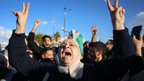 EPA A woman raises both arms and makes the peace sign with her hands in Umayyad Square in Damascus. The sky behind her is a clear blue, and men behind her can be seen celebrating. She looks like she's singing or shouting, with her mouth open and a happy expression.