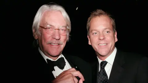 Getty Images Actors Donald Sutherland (l) and Kiefer Sutherland (r) wearing black suits with white shirts. Donald has white hair and glasses and a moustache; Kiefer is clean-shaven with cropped hair and is looking down, both are smiling 