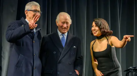 PA Media The King standing with Apple boss Tim Cook, who is making a peace sign, and Raye, who is pointing out into the audience