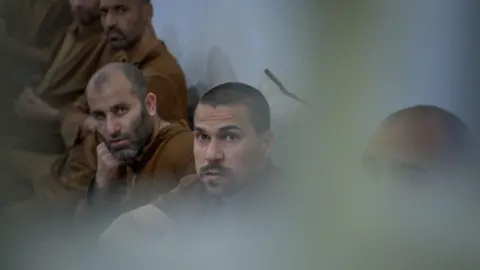 BBC/Michael Steininger Six male inmates sit together in a row against a wall, looking in the same direction, all dressed in the same brown clothing