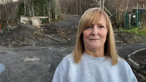 BBC A woman with shoulder length blonde hair and wearing a grey jumper is standing in front of a coal tip. Behind her you can see the area of the coal tip which caused the landslide. There are remains of a shed which was badly damaged and there are also trees surrounding the coal tip area.