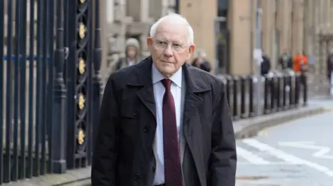 BBC Sir Duncan Nichol, who has short white hair, wearing glasses and a white shirt with a burgundy tie under a black coat, walks towards Liverpool Town Hall. 