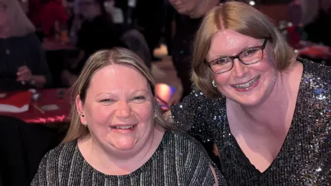 BBC Two women wearing sparkly party tops look towards the camera and smile. Alison, to the left has blond hair and small ear-rings. Laura, to the right, has ginger-blond hair and wears glasses. Tables, decorated for Christmas, can be seen in the background