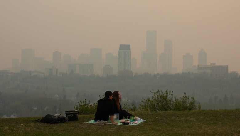 In the foreground, a couple sits on a blanket on grass. A city skyline is visible in the background but a soft orange haze is blanketing everything, making the buildings only partially visible. 