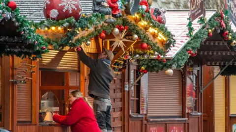 Getty Images A man and a woman work on their stall at a Christmas market in Germany - the woman in a red coat is on the phone while the man tends to his stall