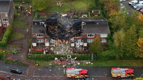 PA Media The house following the explosion. A large crater shaped hole is missing from the rood. Debris from the building is scattered on the nearby grass and  street. Two fire engines are parked on the road and fire fighters are clearing rubble from the building.