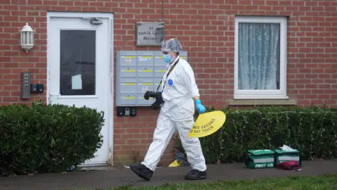 PA Media A forensics officer, in a white suit, hairnet and facemask, is walking from right to left outside terraced housing. The officer has a camera and is holding a yellow cone. There are two green first aid bags on the pavement outside the houses.
