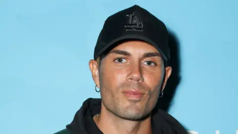 Getty Images Max George wearing a black cap with the letters L and A intertwined. He is stood in front of a light blue background