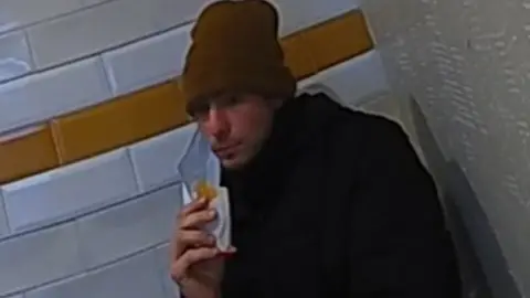 Pennsylvania State Police The suspect, seen eating while seated, in a brown wool hat and dark shirt, with a blue face mask pulled to one side. 