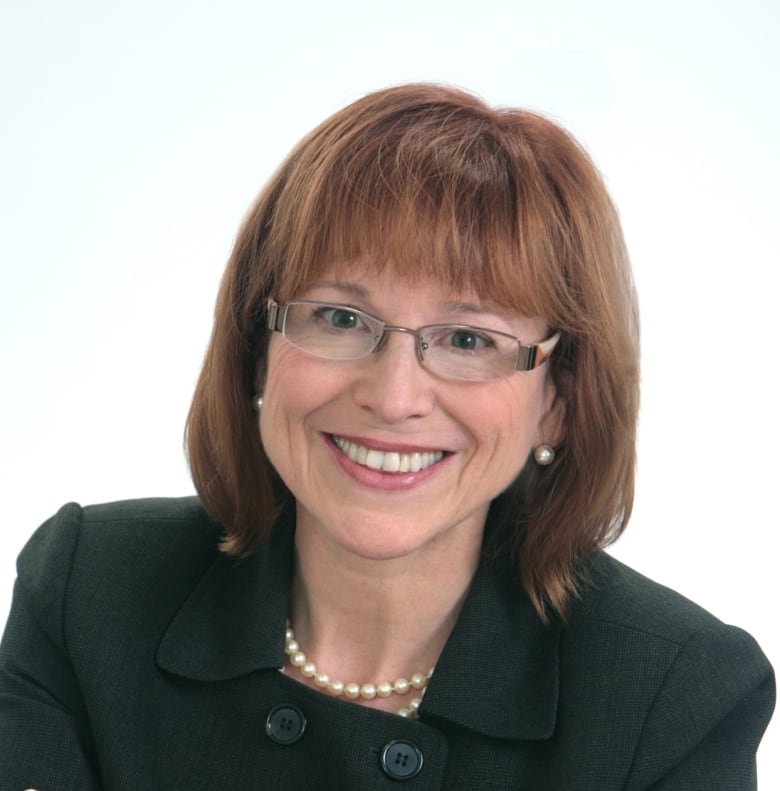 A portrait of a smiling woman with shoulder-length brown hair with bangs, wearing glasses, a black blazer, pearl earrings and necklace.