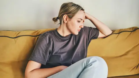 Getty Images A woman wearing a purple T-shirt and light blue jeans sits on a mustard-yellow sofa and turns her face to the side. She has her head resting on her hand and looks unhappy.