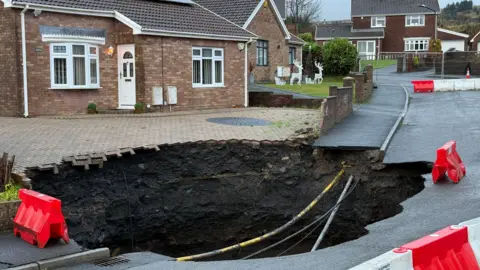 BBC A large and deep hole in the ground can be seen at the end of the driveway of a bungalow. Cables can be seen in the sinkhole and there are a number of barriers in place. The sinkhole includes part of the driveway, the pavement and part of the road. Christmas decorations featuring reindeers and sleighs can be seen in the neighbouring garden.