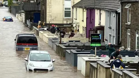 BBC Flooded streets in Sion Street, Pontypridd. Three cars are deep in water in a street that looks more like a river. People wearing raincoats can be seen in the front gardens of terraced houses along the right hand side of the street. There is a pipe over the wall of one of the gardens being used to remove water from a property.