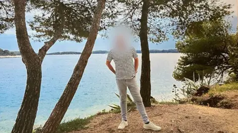 Handout Image shows a man in pale jeans and a striped t shirt. His face has been blurred out. He is stood in front of a lake, surrounded by trees. The sky is blue and he has his hands in his back pockets, looking relaxed.