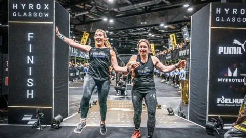 Hyrox and Sportograf Leigh and her friend Wiki are smiling and holding hands as they cross the finish line at an indoor fitness race in Glasgow.