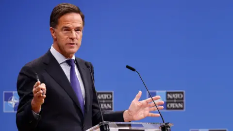 Reuters Mark Rutte wears glasses, a black suit jacket and a purple tie. He speaks into a small microphone on a podium in front of a blue background.