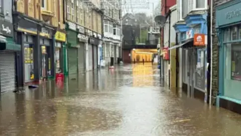 Cerith Mathias Mill Street, with shop fronts on either side and water covering the road