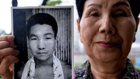 Getty Images Hideko Hakamata wearing a floral shirt holds up a black-and-white photograph of her brother in her right hand, Iwao Hakamata, as a young man