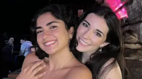 DWAS Two girls smiling at the camera. On the left, the girl has shoulder-length dark hair. She is wearing a blue denim top and silver necklace. The girl on the right has long dark hair. She is wearing a blue denim top and silver earrings. The photo is taken in a bar with long wooden tables in the background.