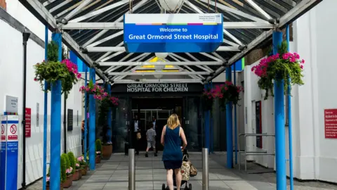 BBC Images A woman with a pushchair is walking into the main entrance of Great Ormond Street Hospital. It's summer and there are baskets of pink flowers hanging from the entrance porch.