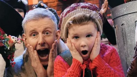 PA Media Patrick Kielty dressed in a Christmas cardigan with children. 
One is dressed as a snowman, one is dressed as a tree and one in a trash can.