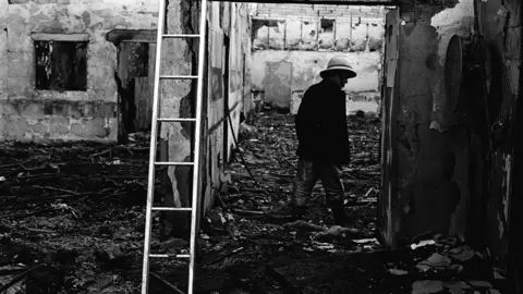 Pacemaker Black and white image of the bombed La Mon hotel, the brick building is covered in black with parts of walls fallen down, debris lies on the floor and a man stands by a ladder in the debris, he is wearing a dark jacket and trousers with wellington boots and a hard hat. 