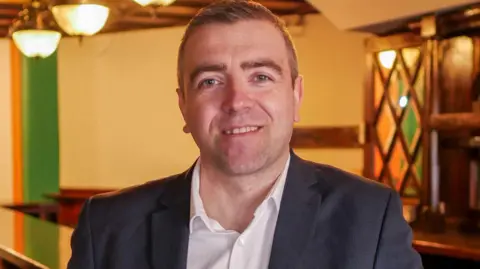 Steven Crosbie A man with short brown hair, a white shirt and a black formal jacket is stood indoors. He is smiling at the camera