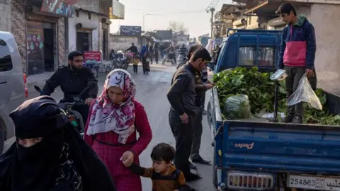 Reuters People walk along a busy street at a refugee camp in Idlib with several people buying vegetables from the back of a truck on 17 December